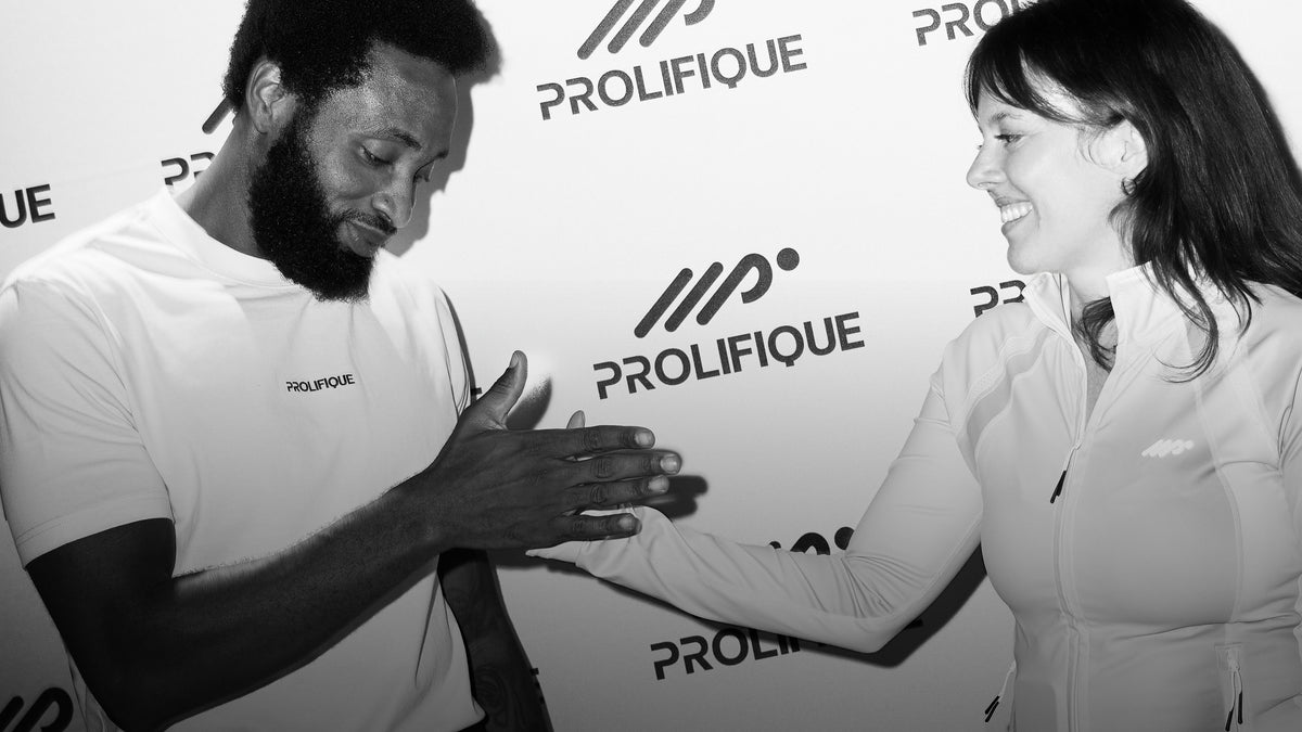 A black and white photo showing a man and a woman shaking hands in front of a branded PROLIFIQUE backdrop. The man wears a green PROLIFIQUE t-shirt, and the woman wears a lime-colored zip-up jacket.