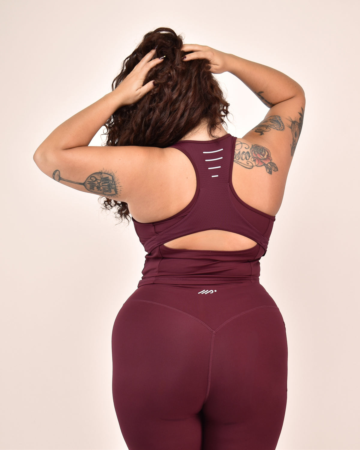 A woman shown from behind wearing a matching maroon activewear set, featuring a sports top with an integrated bra and cutout detail and high-waisted leggings, against a solid light beige background.