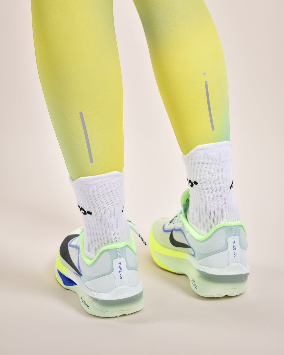 A close-up rear view of a person wearing bright neon yellow-green leggings, white crew socks, and light gray running shoes with vivid neon green and blue accents, set against a solid pale beige background.